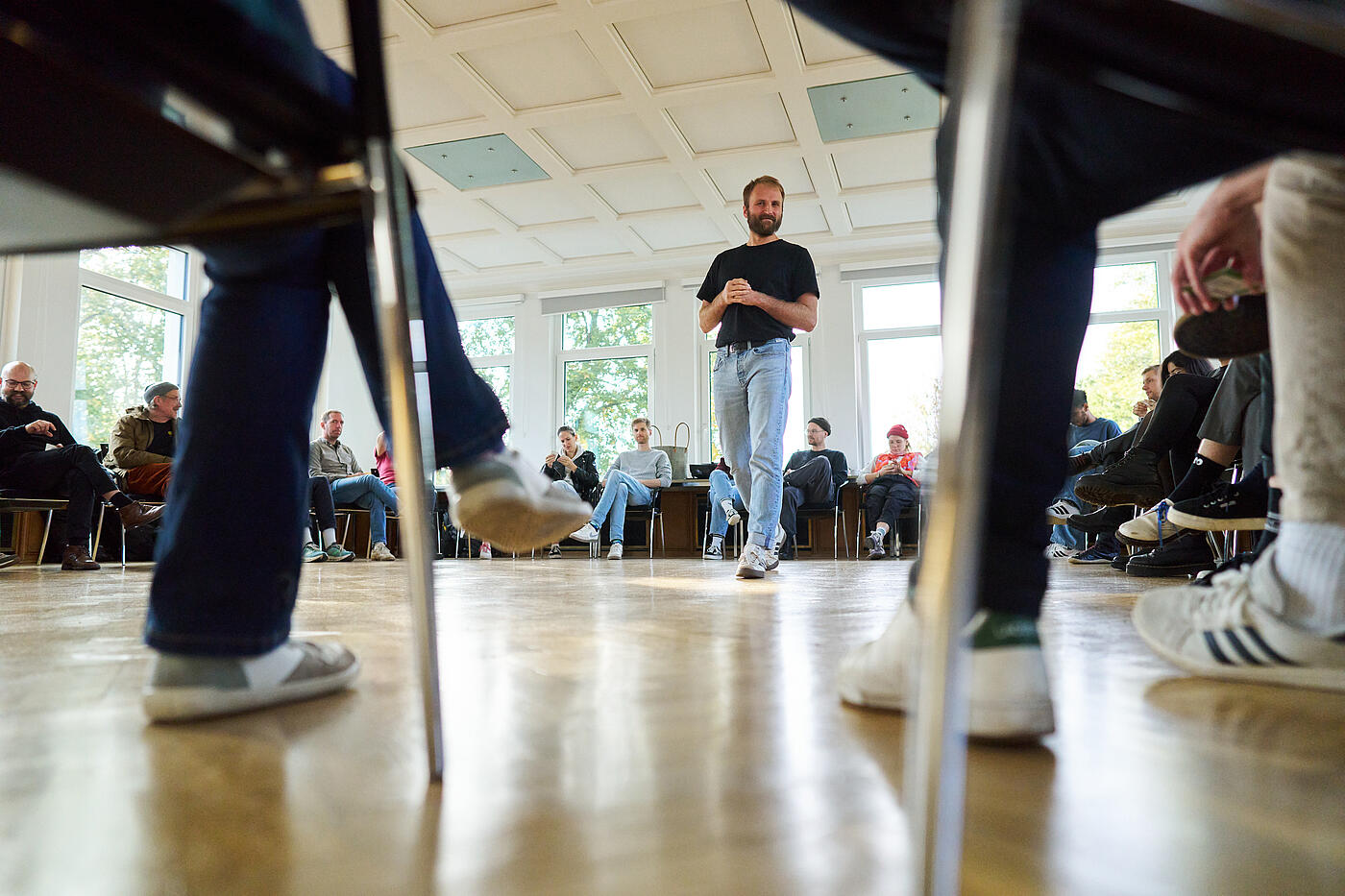 Ein Mann mit Vollbart bewegt sich in blauer Jeans und schwarzem T-Shirt durch die Mitte eines Stuhlkreises.