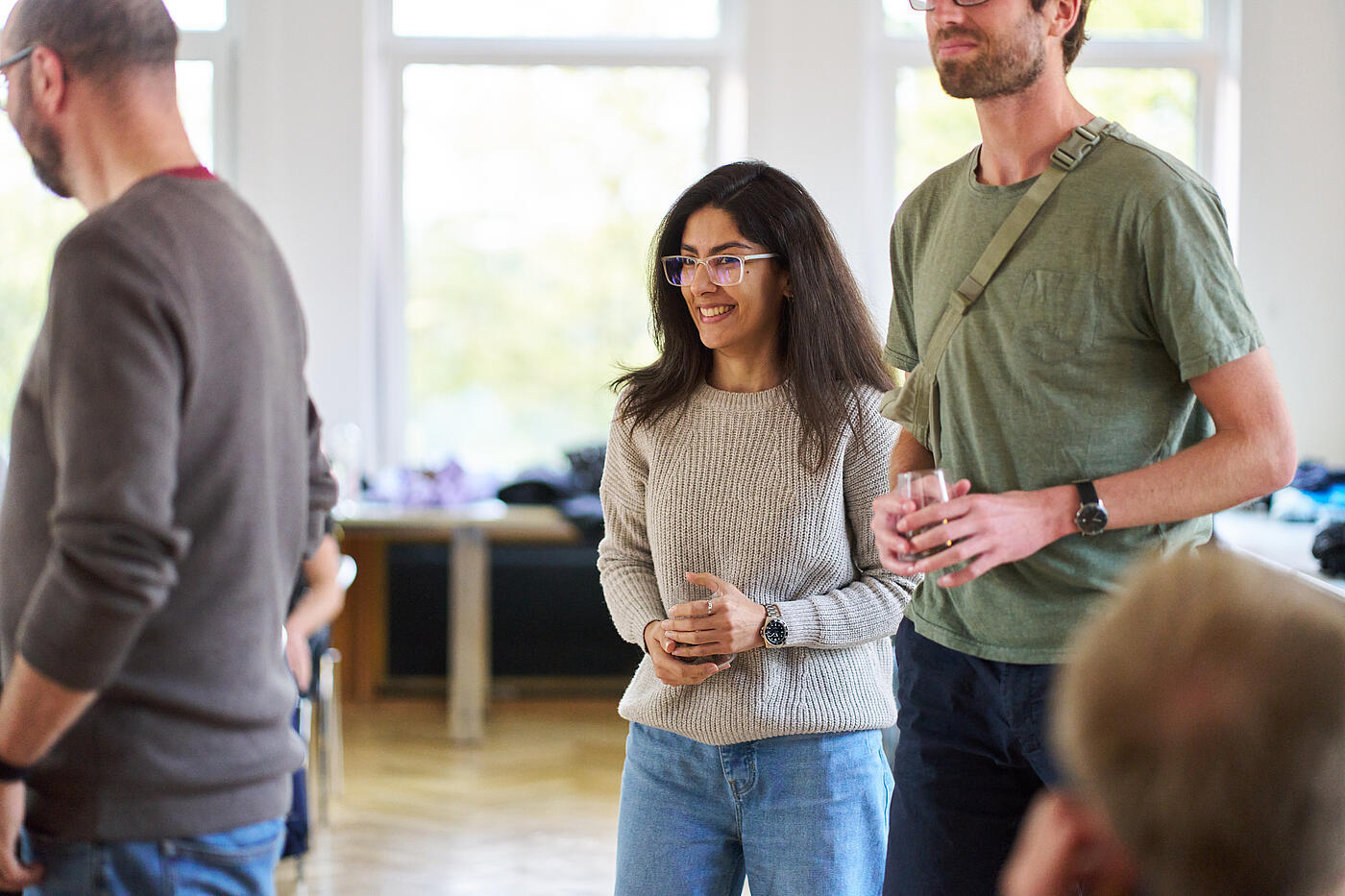 Frau mit dunklen Haaren und Brille in grauem Wollpullover. Neben ihr steht ein Mann in grünem T-Shirt.