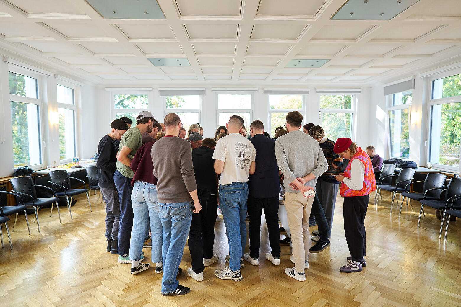 Eine Gruppe Menschen steht in einem Saal mit hellem Parkettboden. Sie bilden einen Kreis und schauen alle in die Mitte.
