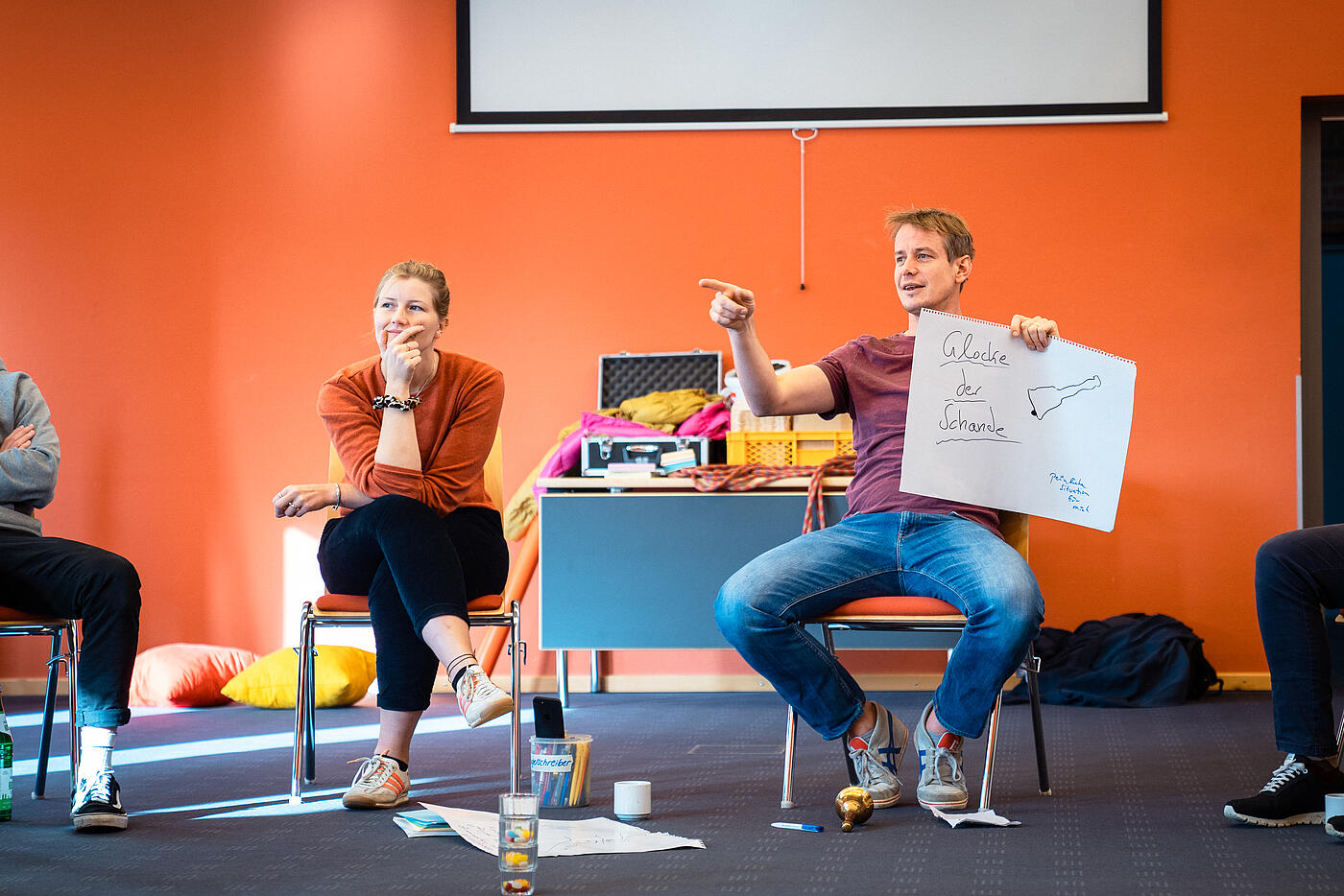 Zwei Personen sitzen vor einer orangen Wand, eine Person hält ein großes Blatt Papier hoch und erklärt etwas.