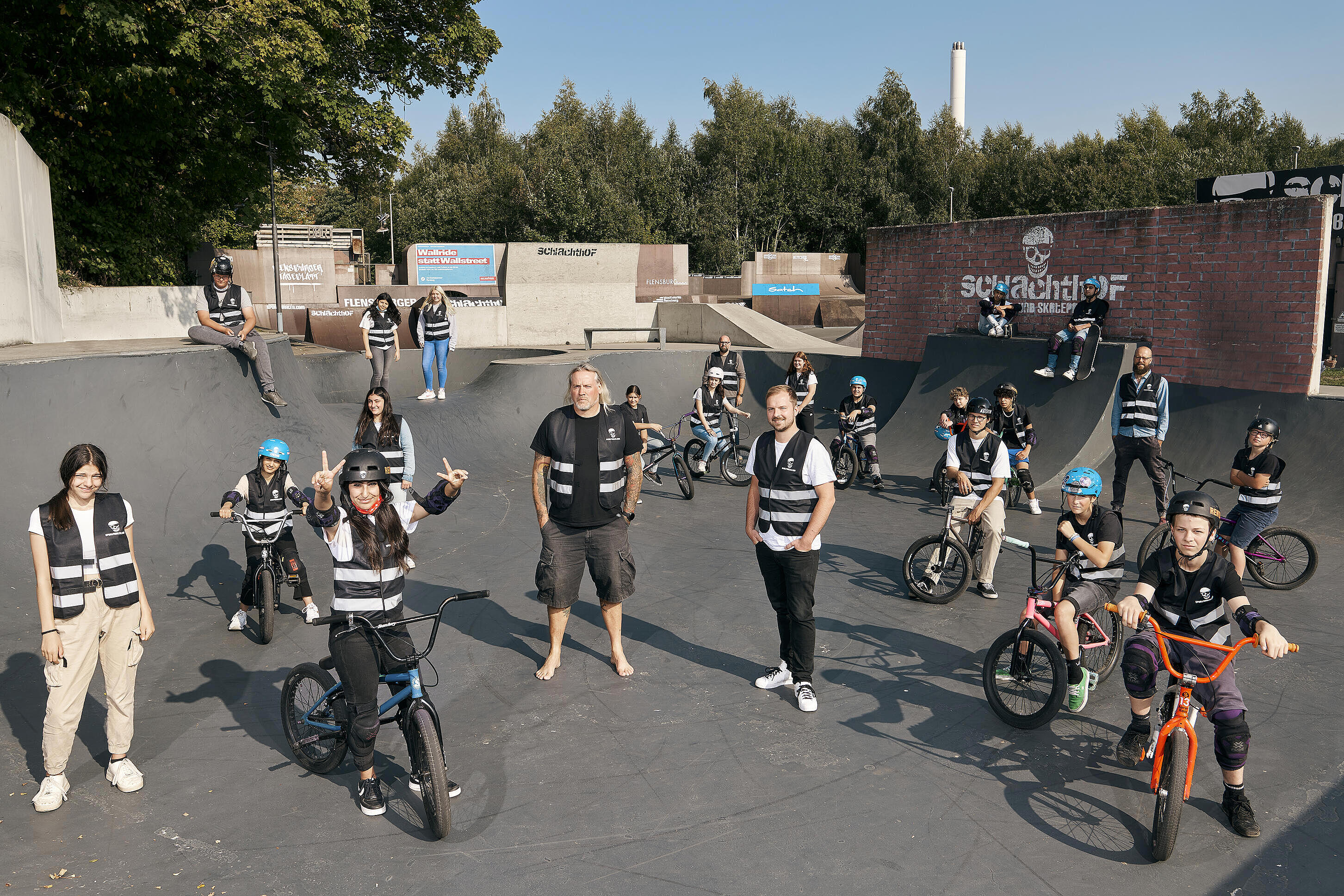 visuellverstehen-Geschäftsführer und Sportpiraten-Chef inmitten junger BMX-Fahrer*innen auf dem Gelände der Sportpiraten. Alle Personen tragen die schwarzweiße Warnweste mit Sportpiraten- und visuellverstehen-Aufdruck.