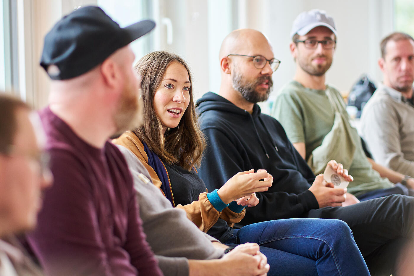 Eine Frau mit dunklen Haaren sitzt auf einem Stuhl und streckt die rechte Hand hervor. Neben ihr sitzen Männer auf Stühlen.