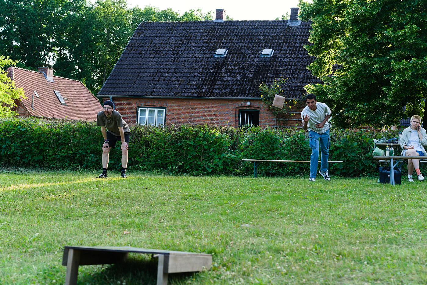 Zwei Mitarbeiter spielen auf einer Grünfläche Cornhole, im Hintergrund eine Hecke und ein historisches Gebäude.