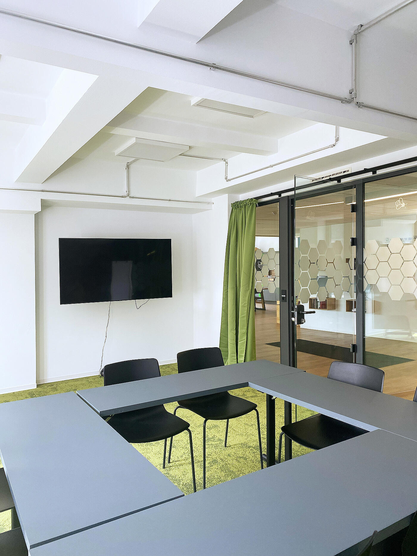 Kleiner Besprechungsraum mit vier Tischen, die zu einem Quadrat mit Loch in der Mitte zusammengestellt sind. Teppich und Gardine sind grün, es hängt ein großer Screen an der Wand.