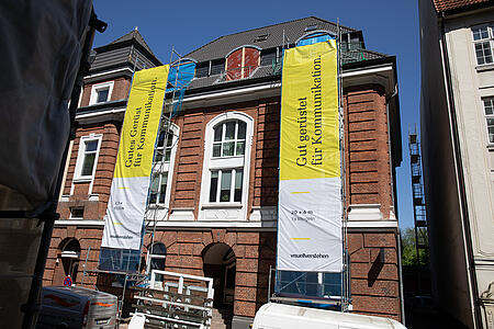 Zwei große gelbe visuellverstehen-Banner hängen an den Gerüsten vorm Agenturgebäude. 