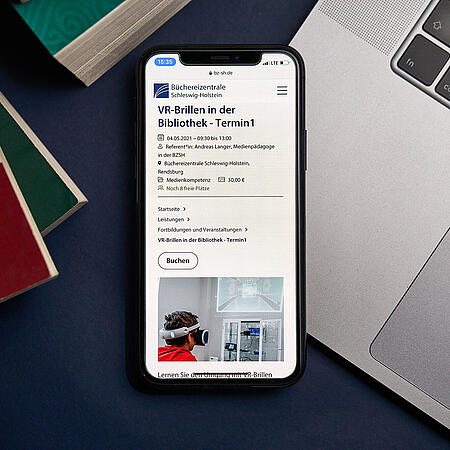 Ein Smartphone, das auf einem Macbook liegt, zeigt den neuen Internetauftritt der Büchereizentrale SH inmitten von Bücherstapeln auf einem Tisch.