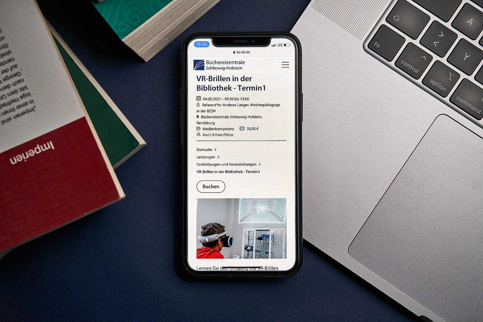 Ein Smartphone, das auf einem Macbook liegt, zeigt den neuen Internetauftritt der Büchereizentrale SH inmitten von Bücherstapeln auf einem Tisch.