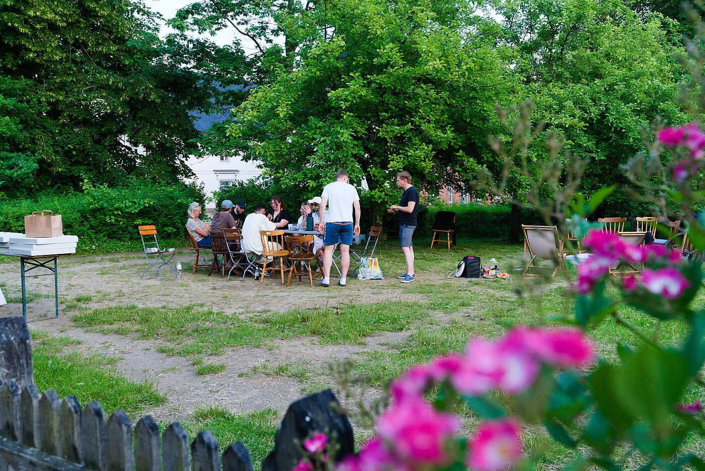 Vor einem idyllischen Hintergrund mit Bäumen und einem historischen Gebäude sitzen mehrere Teammitglieder an einem Tisch im Schatten. Im Vordergrund rahmen Rosen das Bild.