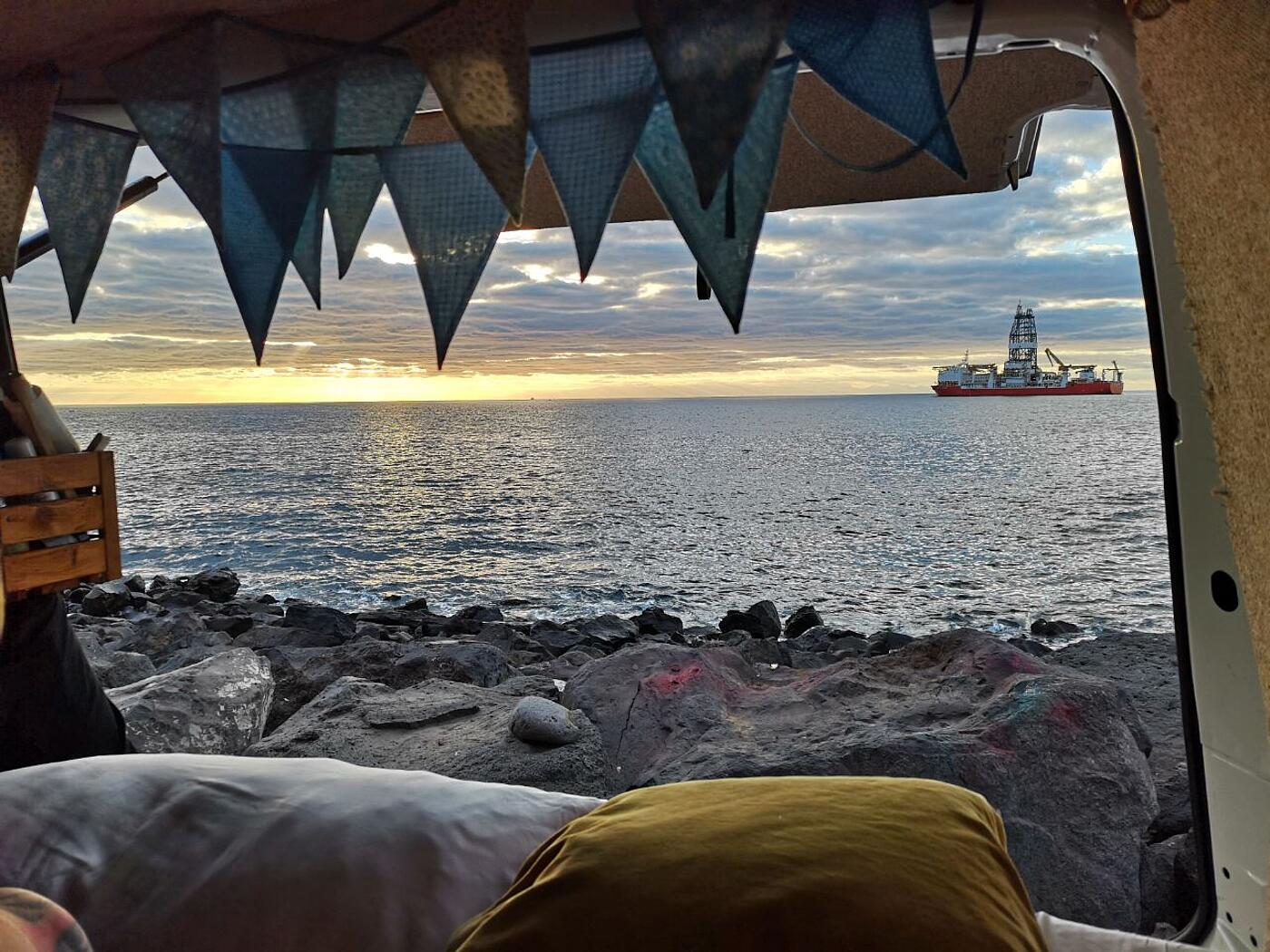 Der Blick aus dem Inneren des VW-Busses durch die Heckklappe hinaus, dahinter Strand, Meer und ein großer Dampfer.