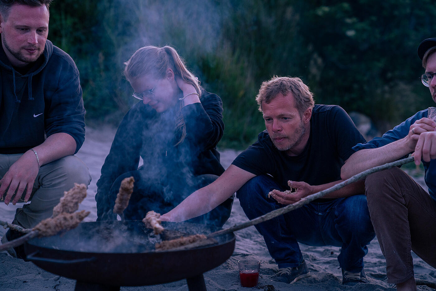 Timon sitzt mit drei weiteren Personen an einer Feuerschale. In der Hand hält er einen Stock mit Brotteig.