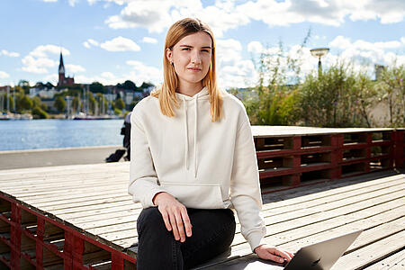 Eine Mitarbeiterin von visuellverstehen sitzt am Flensburger Hafen in der Sonne.