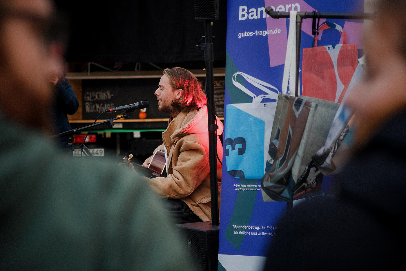 Ein Musiker in brauner Lederjacke sitzt mit Gitarre vor einem RollUp zu den Bannerbeuteln von visuellverstehen.