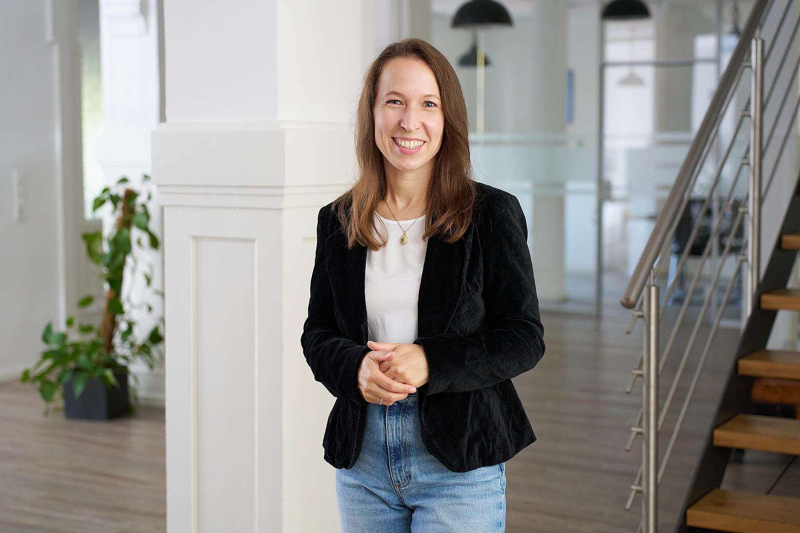Anna steht lächelnd in den Büroräumen von visuellverstehen. Sie trägt langes braunes Haar, ein weißes T-Shirt und einen dunklen Blazer.