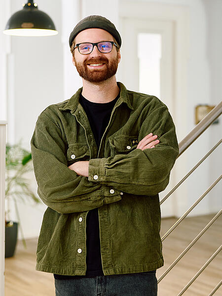 Lukas lehnt lächelnd an einer Treppe in den Büroräumen von visuellverstehen. Er trägt einen braunen Vollbart, ein Beanie, eine Brille und eine grüne Cordjacke.