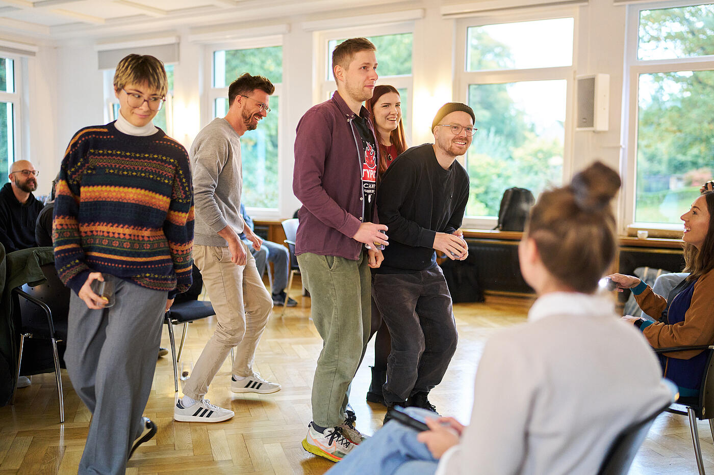 Zwei Frauen und drei Männer stehen mit gebeugten Knien und teilweise gesenkten Köpfen. Die meisten lachen und halten ein Glas in der Hand.