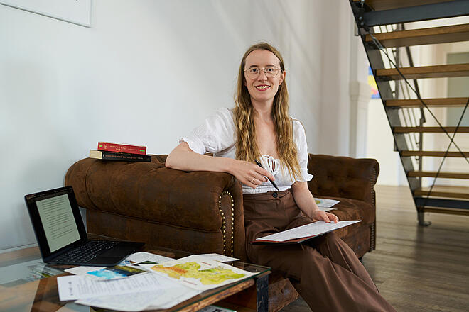 Ragna auf einem braunen Sofa mit einem Papier auf dem Schoß. Sie hat lange, braune Haare, trägt eine Brille sowie ein weißes Oberteil und eine braune Hose. Auf dem Tisch neben ihr liegen Papiere und ein aufgeklapptes MacBook.