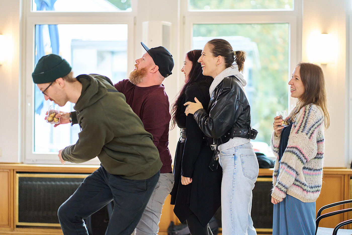 Zwei Männer mit Kopfbedeckungen im Ungleichgewicht. Daneben stehen drei Frauen und lachen.