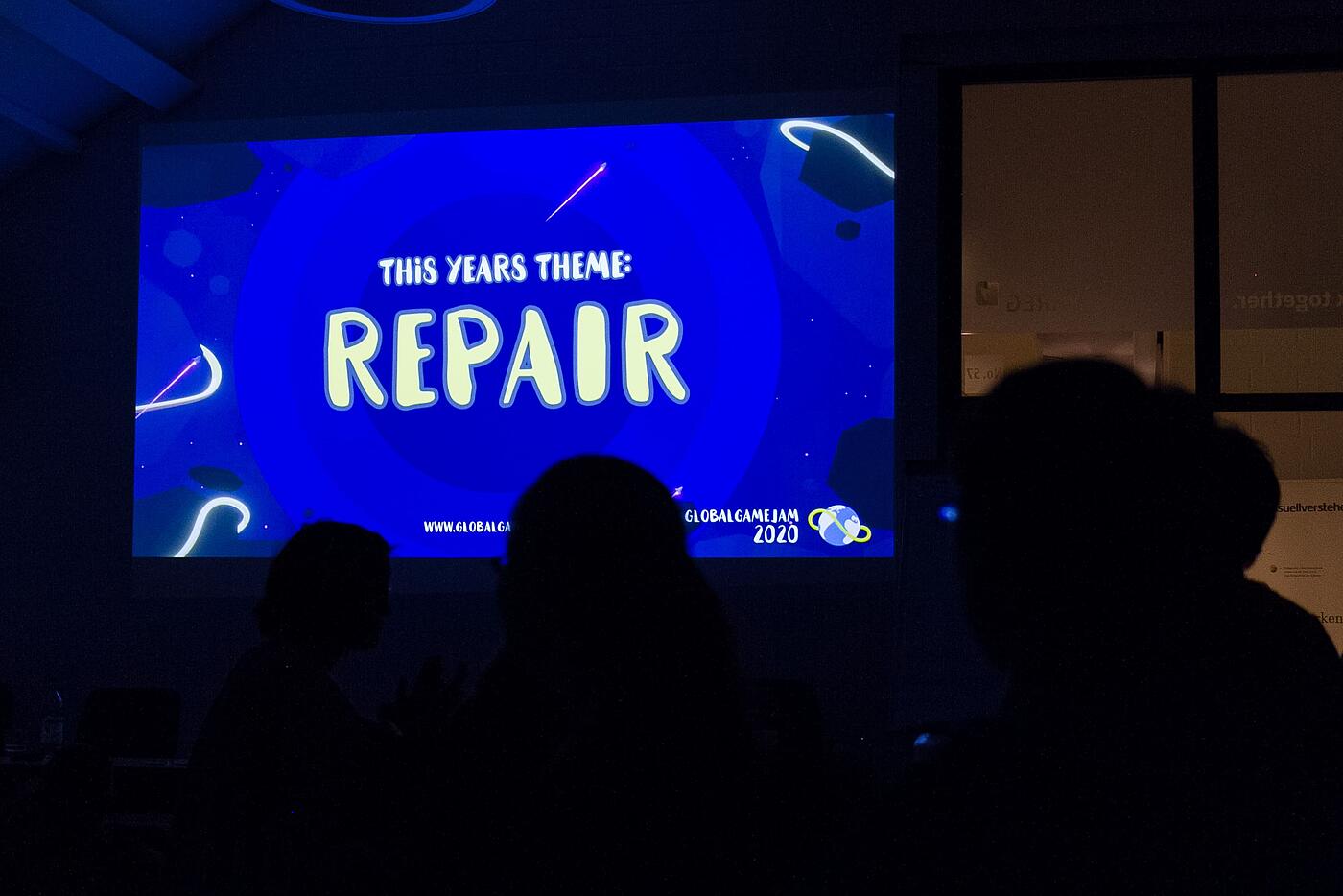 Auf dem Präsentationsbildschirm bei einer Konferenz steht auf blauem Hintergrund, dass das diesjährige Thema Repair ist.