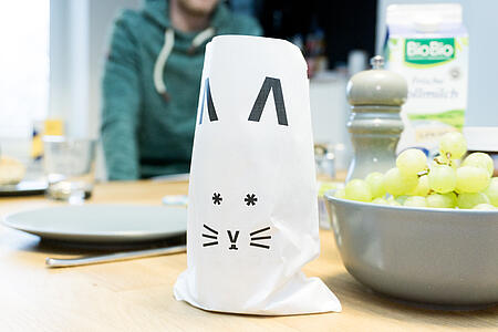 Auf einem Tisch stehen eine Obstschale und eine Papiertüte, auf die das Gesicht eines Osterhasen aufgedruckt ist.