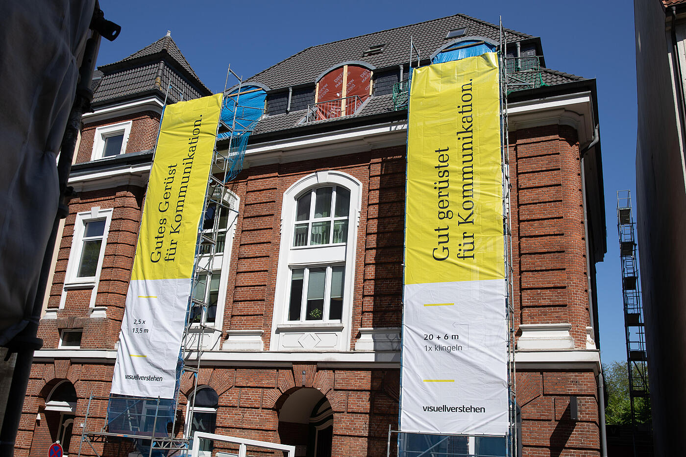 Vor dem visuellverstehen-Gebäude hängen zwei riesige gelbe visuellverstehen-Banner von Baugerüsten herab.
