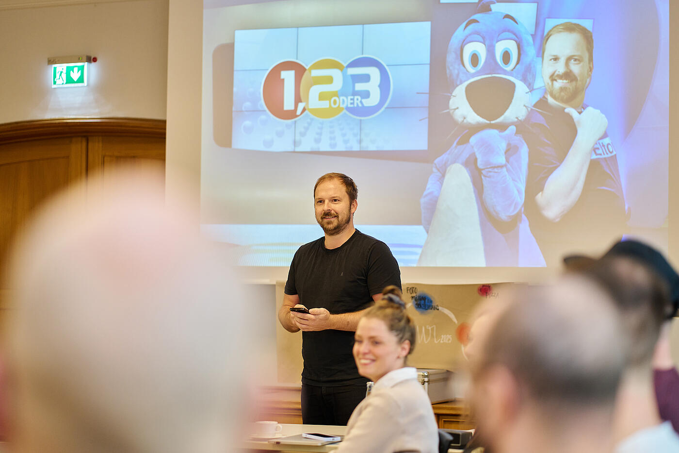 Ein Mann in schwarzem T-Shirt steht vor einer Leinwand. Auf der Leinwand steht 1, 2 oder 3. Daneben eine blaue Robbe und ein Foto des Mannes mit gehobenem Daumen.