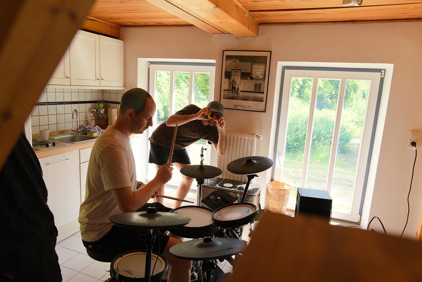 Ein Mitarbeiter spielt in einem warm erleuchteten Innenraum Schlagzeug an einem E-Drum-Set. Im Hintergrund fotografiert ein weiterer Mitarbeiter vorgebeugt den Fotografen dieses Bilds mit einer Einwegkamera.