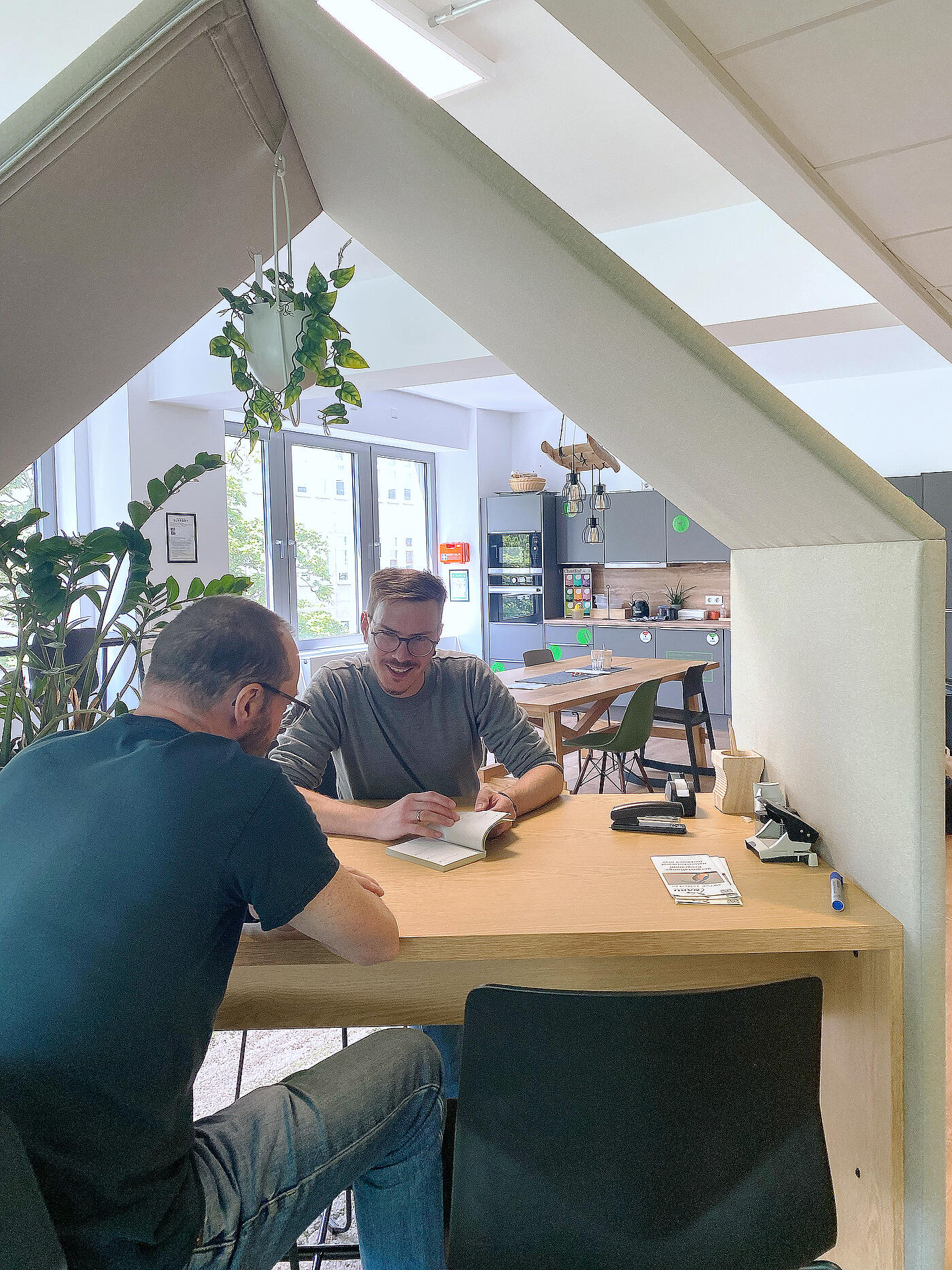 Zwei Männer sitzen sich an einem Schreibtisch gegenüber. Im Hintergrund ist eine offene Küche sowie große Fenster.