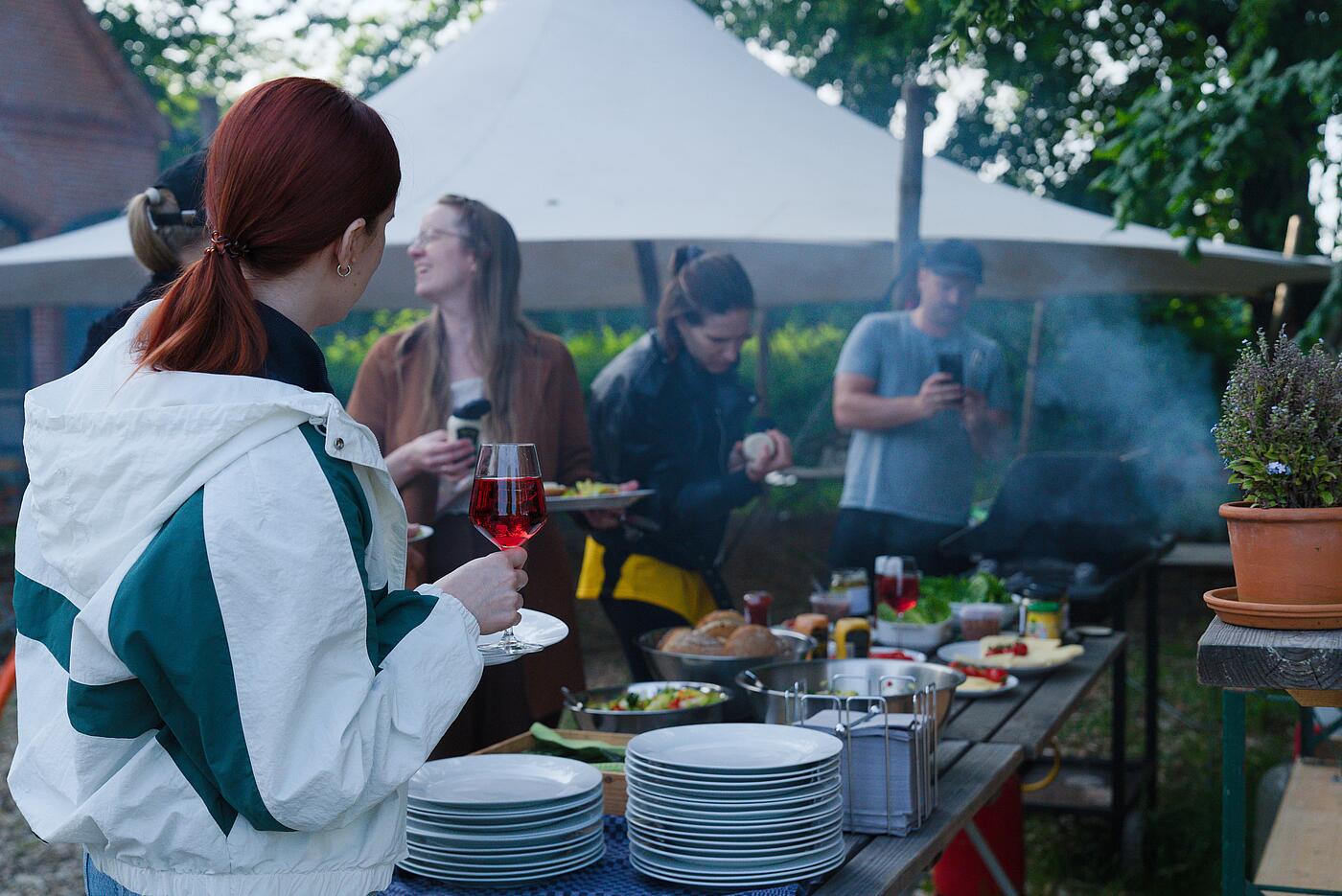 Mehrere Teammitglieder stehen an an einem Buffet für Burger, im Hintergrund ein Pavillon und Bäume, Rauch zieht durchs Bild. Eine Mitarbeiterin lacht lauthals.