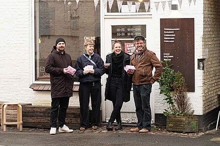 Vier Personen stehen lachend nebeneinander vor der weißen Backsteinfassade eines Cafés mit dem Namen „ONOMA“. Sie halten kleine, rosafarbene Päckchen in den Händen. Über dem Eingang hängt eine Wimpelkette, rechts im Bild ist eine Durchfahrt mit weiteren Sitzgelegenheiten zu sehen.