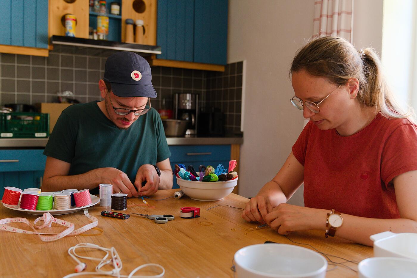 Zwei Mitarbeitende sitzen in einem Innenraum an einem Tisch und knüpfen konzentriert Armbänder. Auf dem Tisch stehen Porzellanschalen mit Materialien.
