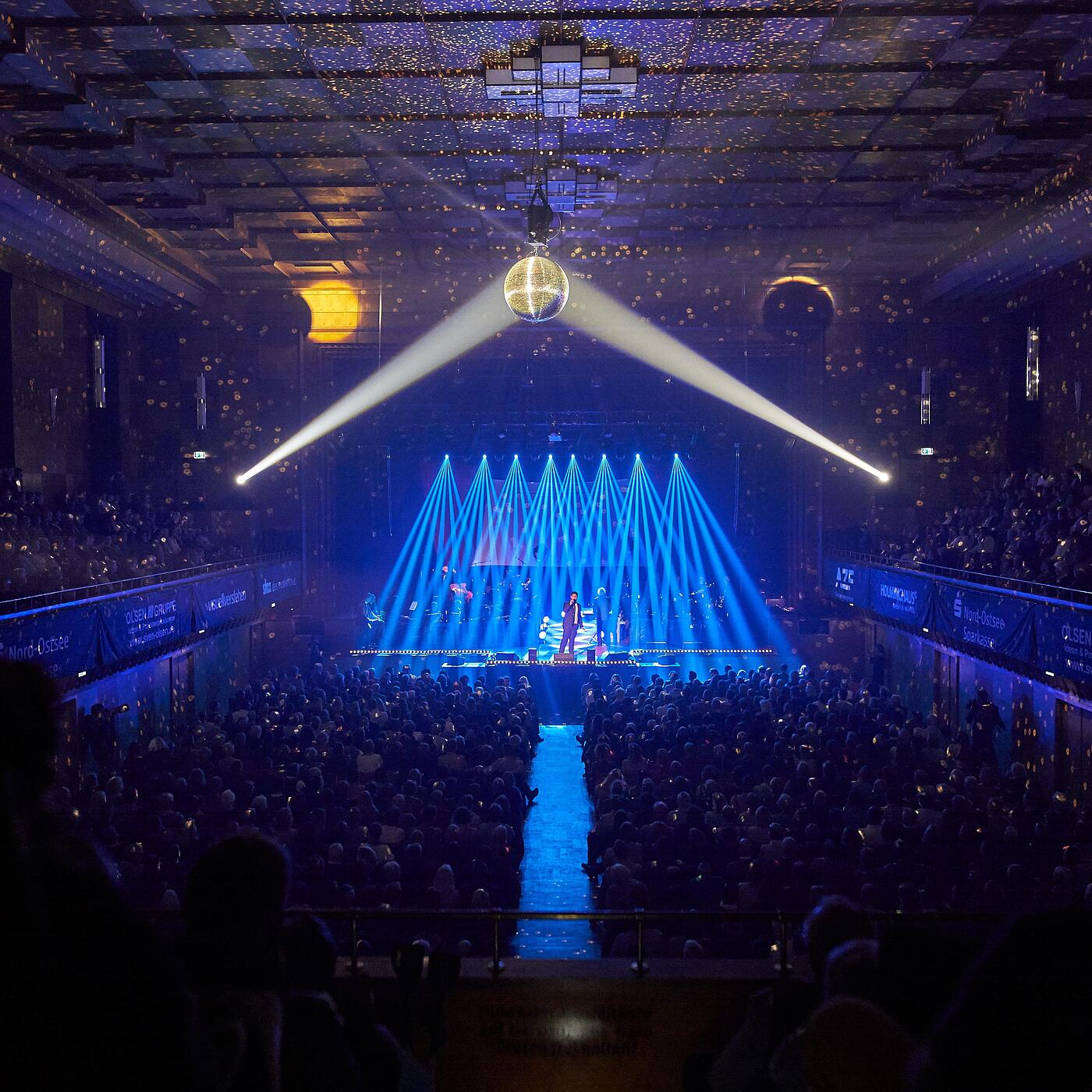 Person singt vor großem Publikum auf der Flensburg-singt-Sinatra-Veranstaltung, gehüllt in blaues Licht, aus einer etwas anderen Perspektive.