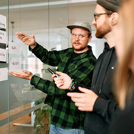 Drei Mitarbeitende von visuellverstehen, die ein Projekt mit Statty Notes an einer Glaswand visualisieren und sich darüber austauschen.