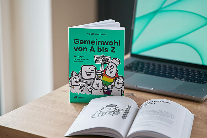 Das visuellverstehen-Buch Gemeinwohl von A-Z steht neben einem Macbook und einem aufgeschlagenen Exemplar desselben Buchs auf einem Tisch.