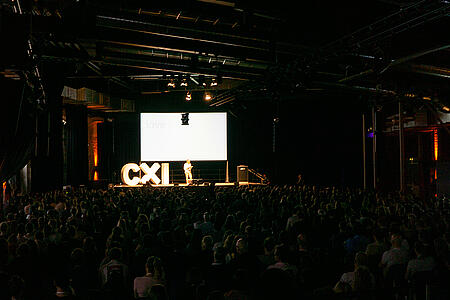 Ein vollbesetzter, dunkler Saal mit einer hell erleuchteten Bühne bei der CXI-Konferenz in Berlin.