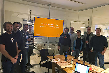 Das Team von visuellverstehen posiert in einem verglasten Besprechungsraum vor einem orangen Bildschirm mit einer Typo3-Überschrift.