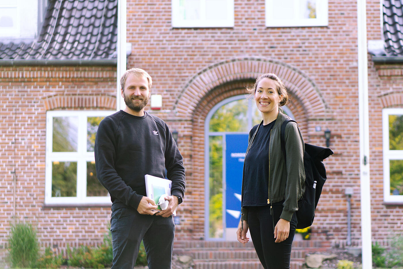 Malte Riechmann und Frauke Hellwig stehen vorm Eingang einer der Bildungsstätten und lächeln in die Kamera.