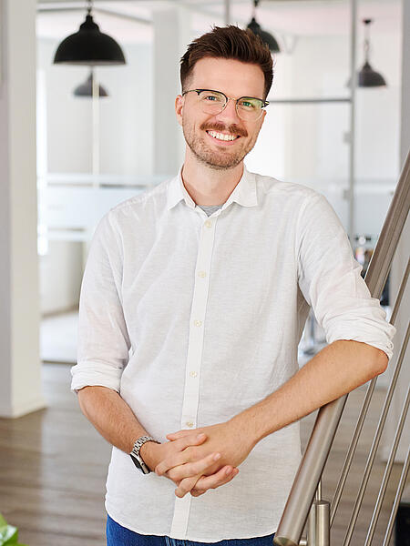 Lars steht lächelnd neben einer Treppe in den Büroräumen von visuellverstehen. Er trägt kurzes, braunes Haar, eine Brille und ein weißes Hemd.