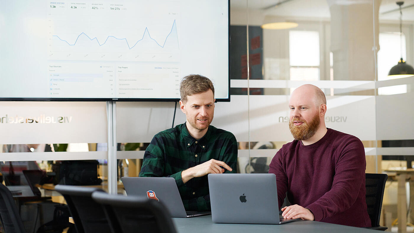 Zwei Mitarbeiter von visuellverstehen sitzen vor ihren Macbooks und besprechen etwas.