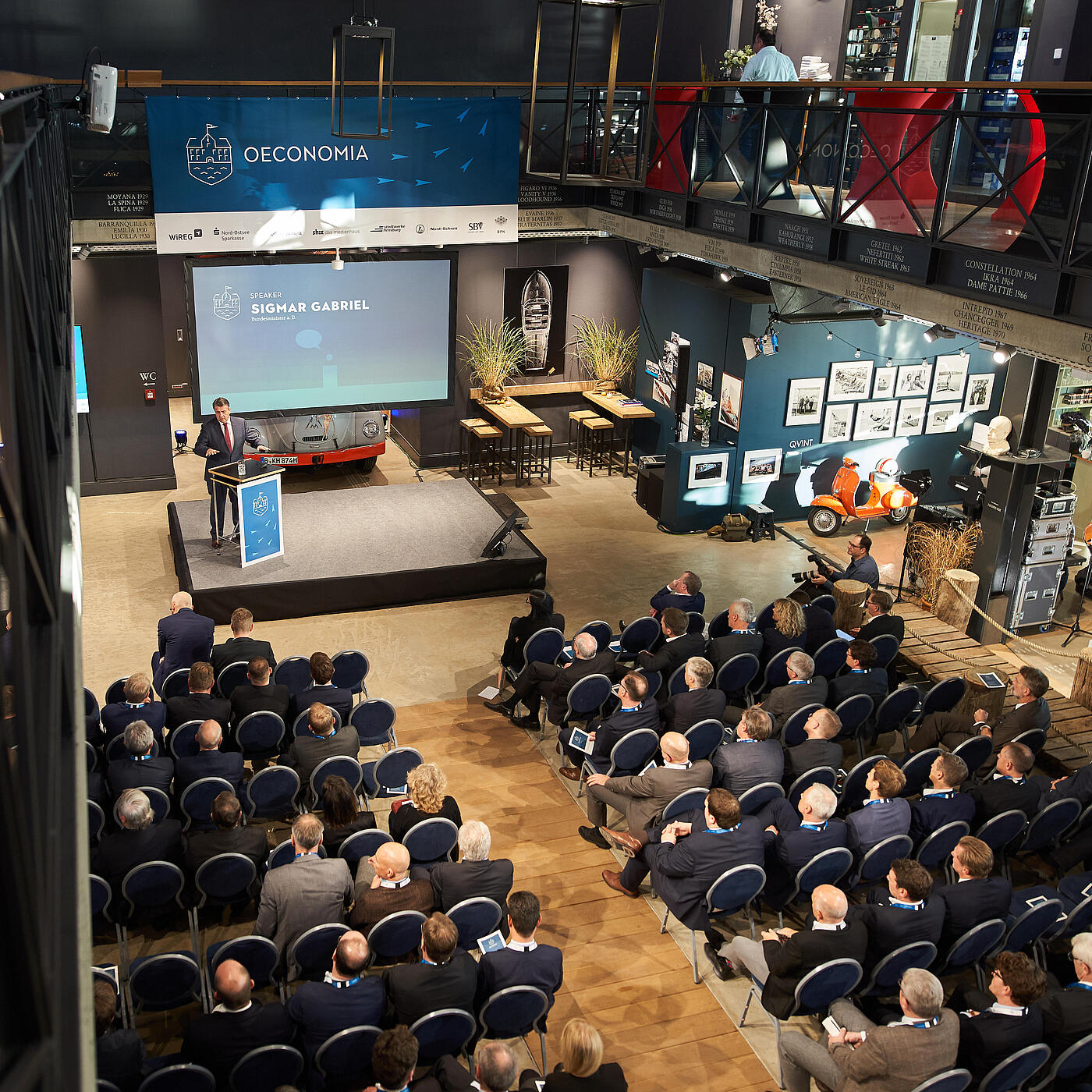 Ein Blick aus der Vogelperspektive auf die Oeconomia 2018, bei der viele Menschen in einem Saal sitzen und vorn zur Bühne schauen.