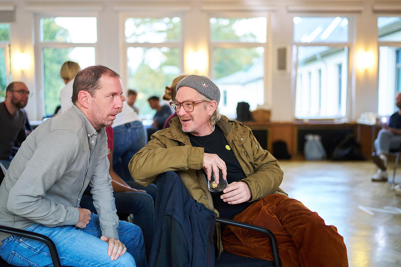 Zwei Männer sitzen hintereinander auf Stühlen. Der vordere hat sich zum Hintermann hin umgedreht und lächelt.