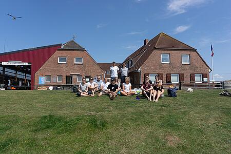 Das Team von visuellverstehen sitzt im Gras in der Sonne, im Hintergrund zwei Gebäude.
