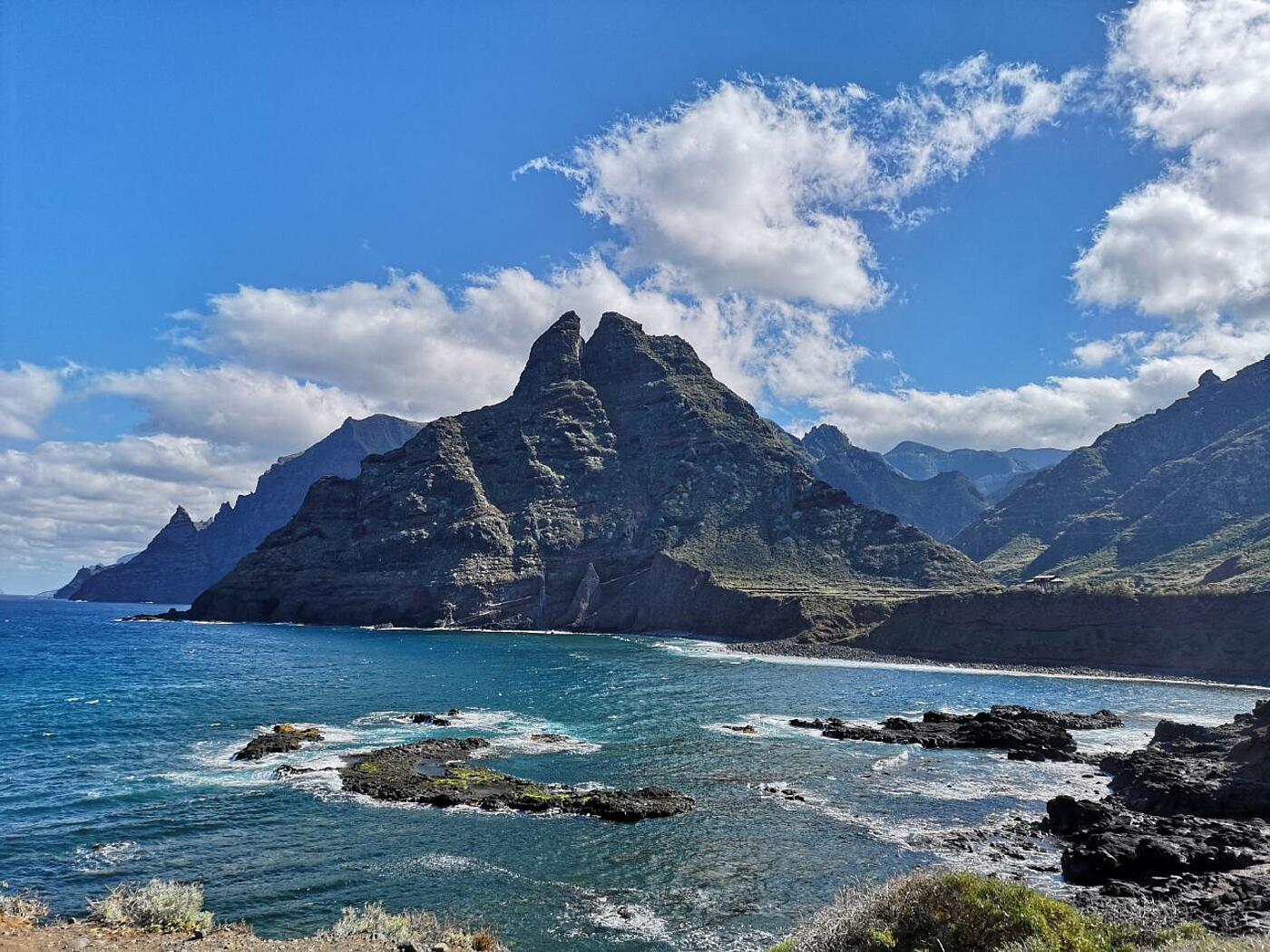 Eine ansprechende Landschaft mit Meer im Vordergrund und schroffen Felsen im Hintergrund, die Sonne scheint.