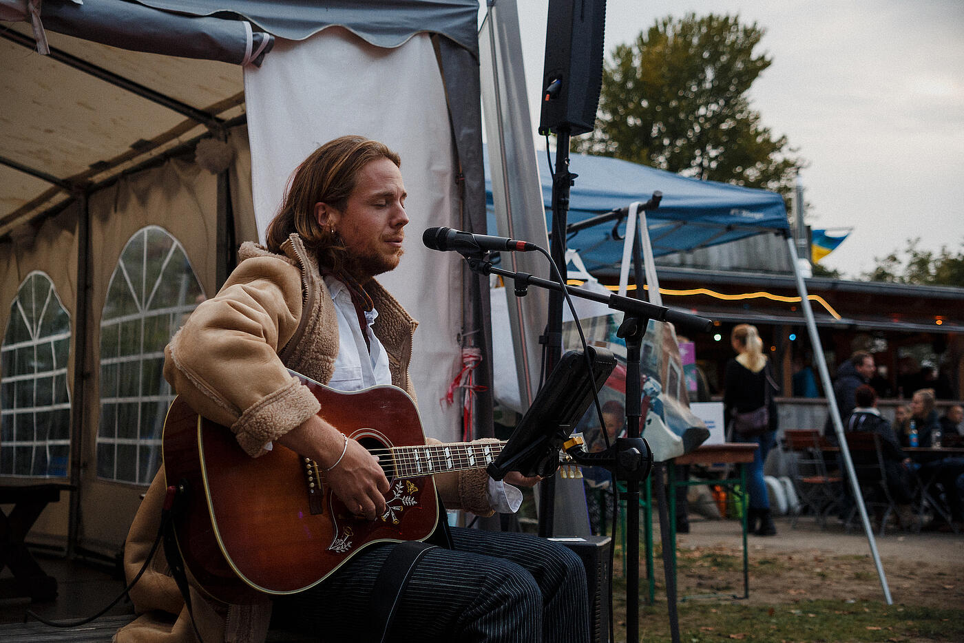 Ein Musiker in brauner Lederjacke sitzt mit Gitarre vor einem Zelt und singt in ein Mikrofon.