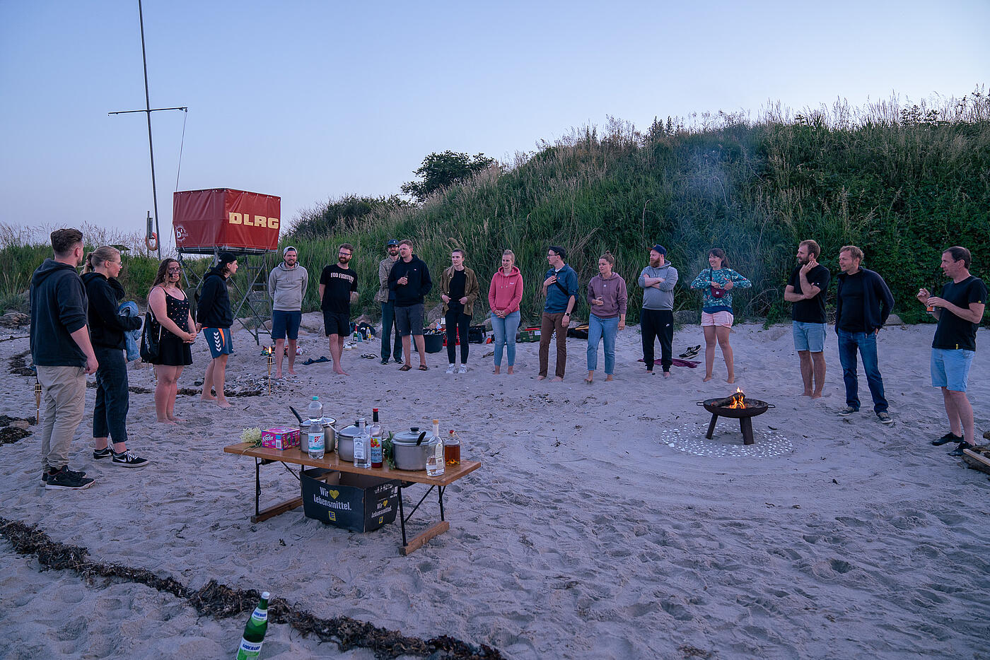Mitarbeitende von visuellverstehen stehen am Strand in einem großen Kreis. In der Mitte brennt ein Lagerfeuer.