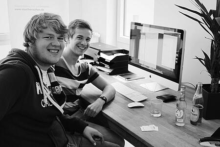 Zwei junge Mitarbeiter von visuellverstehen sitzen am Arbeitsplatz und lachen in die Kamera.