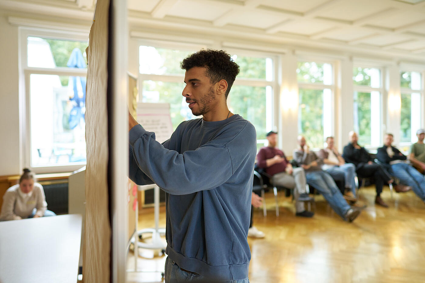 Junger Mann mit dunklen gelockten Haaren und blauem Sweatshirt steckt etwas an eine Pinnwand.