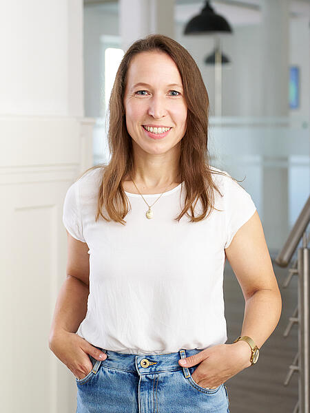 Anna steht lächelnd in den Büroräumen von visuellverstehen. Sie trägt langes braunes Haar, ein weißes T-Shirt und helle Jeans.