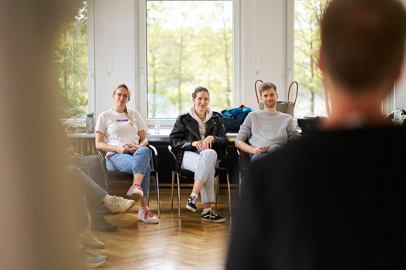 Zwei Frauen und ein Mann sitzen nebeneinander auf Stühlen vor einem Fenster. Auf ihren Namensschildern steht Julia, Cleo, Moritz.