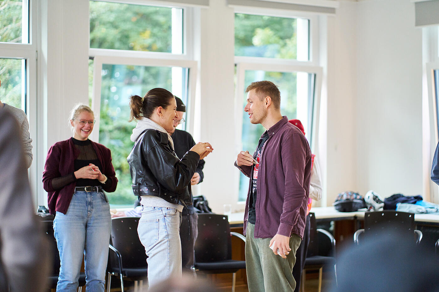 Eine Frau und ein Mann stehen sich gegenüber und unterhalten sich. Im Hintergrund weitere Personen und Fenster.