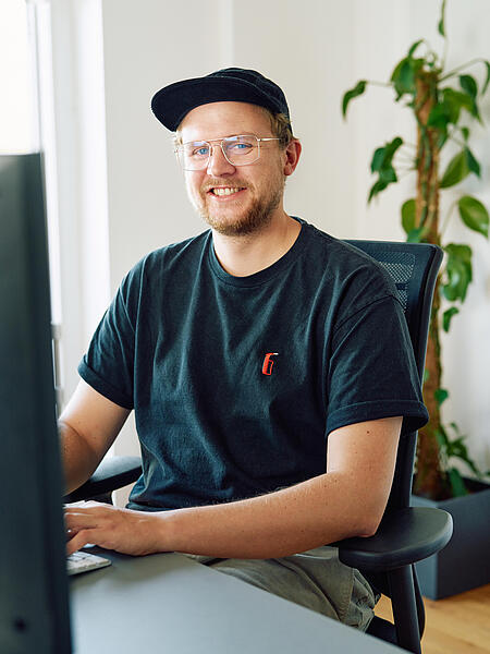 Timon sitzt lächelnd an einem Schreibtisch in den Büroräumen von visuellverstehen. Er trägt einen kurzen Bart, ein schwarzes Cap, eine Brille und ein schwarzes T-Shirt. 